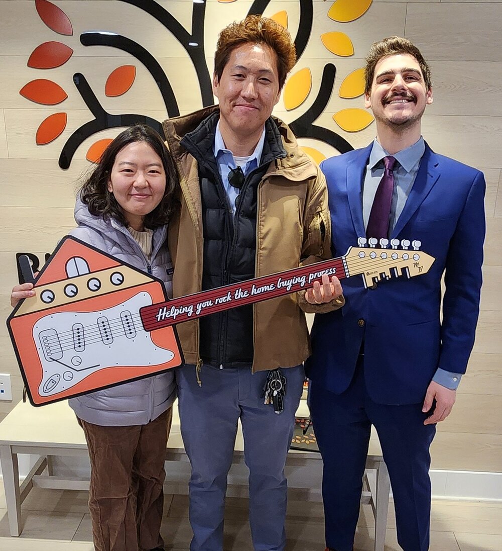 Michael Basch with happy clients at their home closing, holding the Mike Basch Mortgage Team guitar sign